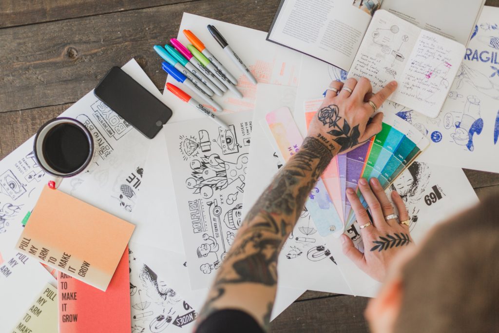 Em uma mesa de madeira um designer espalhou várias folhas de papel desenhadas, canetas e está escolhendo uma cor em uma cartela de cores. O design de uma marca está em todos os detalhes. 