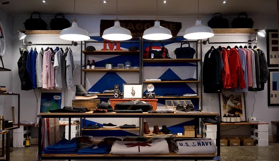 Foto interna de uma loja de roupas masculinas, com uma ilha de acessórios ao centro e araras com camisas coloridas ao fundo.