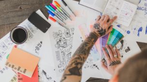 Em uma mesa de madeira um designer espalhou várias folhas de papel desenhadas, canetas e está escolhendo uma cor em uma cartela de cores